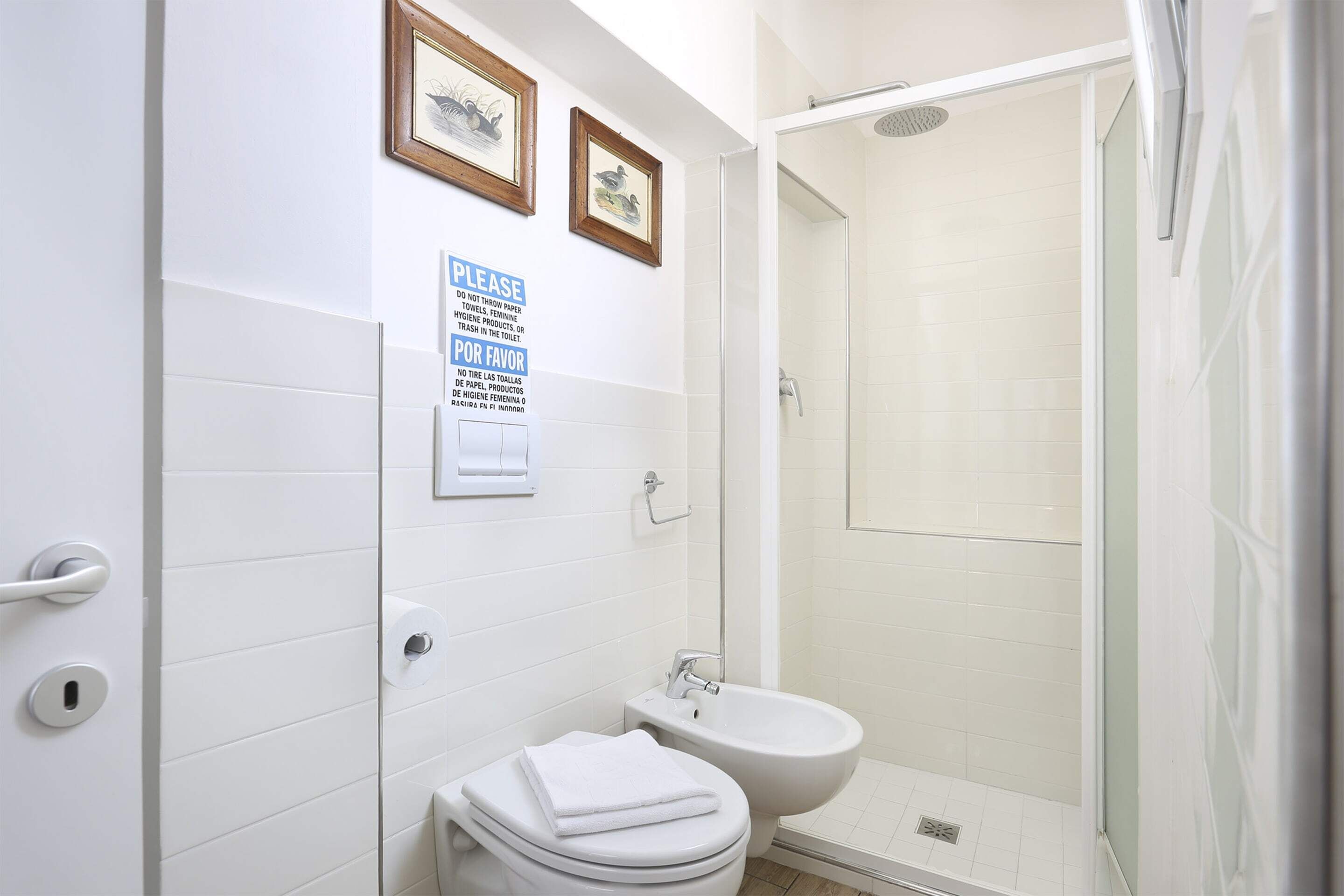 Une salle de bain équipée de toilettes, d’un bidet et d’une douche à parois de verre. Deux tableaux encadrés ornent le mur, ainsi qu’un panneau indiquant les consignes d’hygiène.