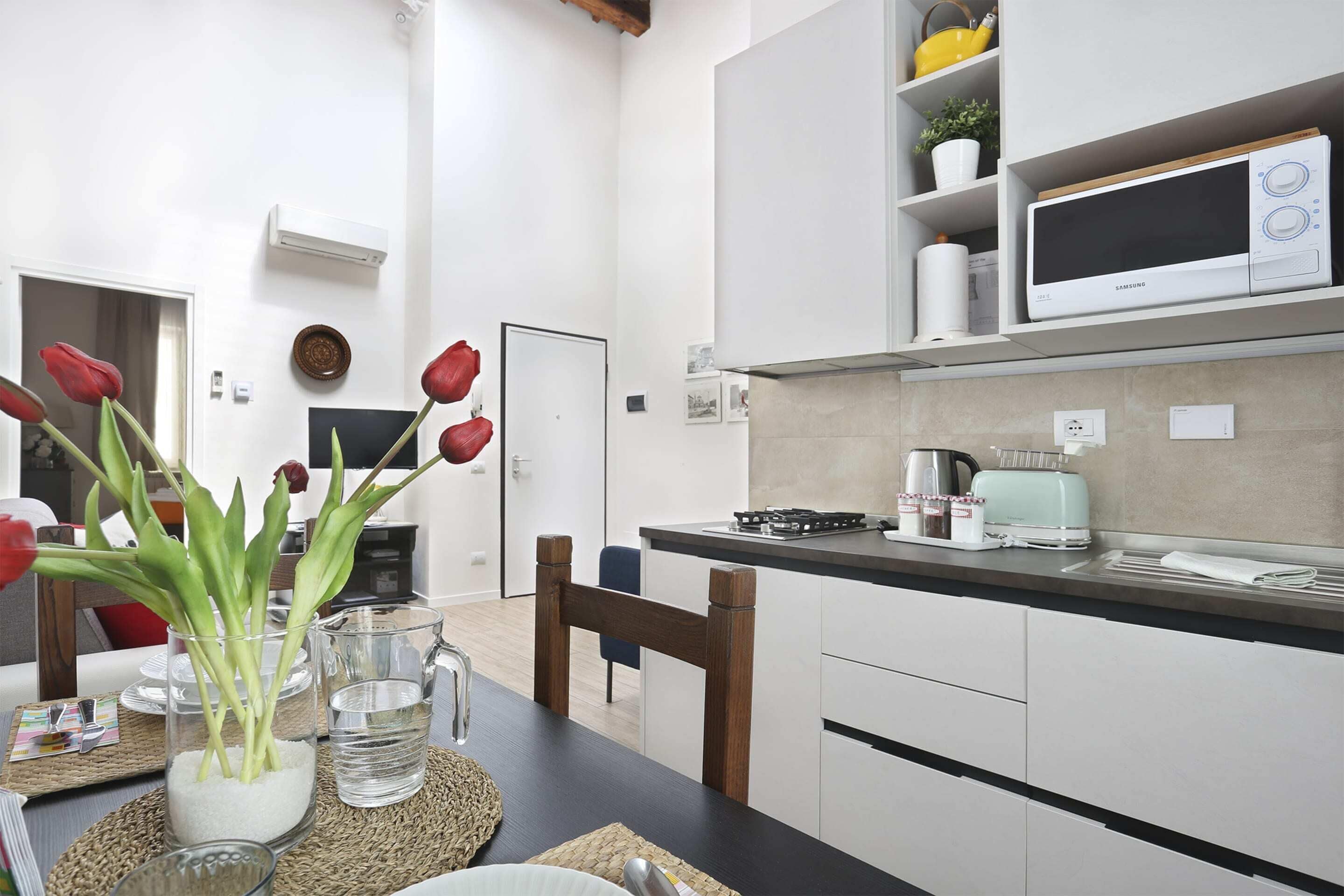 Una cocina moderna con armarios blancos, microondas y tostadora. En la encimera hay un jarrón con tulipanes rojos.