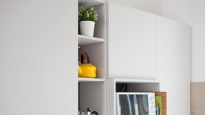 Un mobile bianco con un forno a microonde e un bollitore sullo scaffale inferiore, un peperone giallo sullo scaffale centrale e una pianta in vaso sullo scaffale superiore.