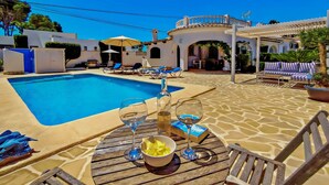 Zona de piscina amb gandules, una taula amb acompanyaments i una pèrgola.