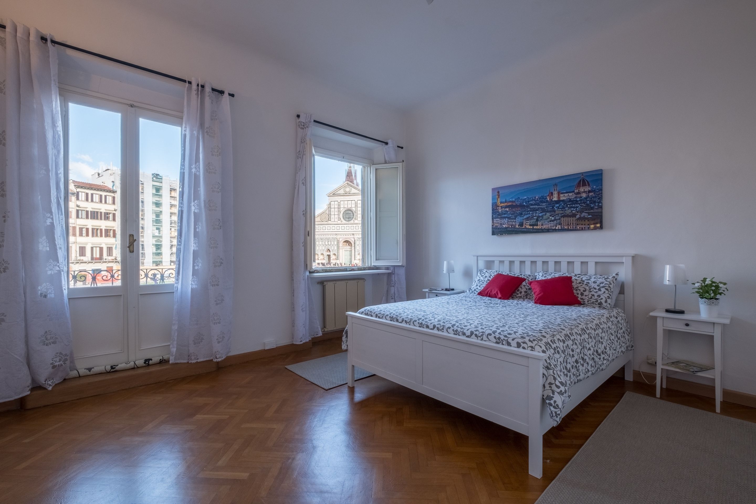 A bedroom with a bed, a window, a painting, and a nightstand.