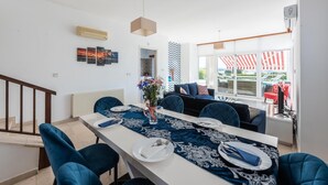 Spiseområde med et langt bord dækket til fire personer, blå stole, en vase med blomster og en moderne pendellampe.
