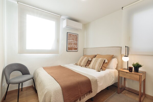 Un dormitorio con cama, una silla, una mesita de noche con lámpara y una ventana.