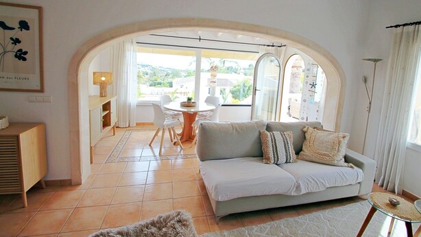 A living room with a sofa, a coffee table, and a view through an arched doorway to an outdoor area with a table and chairs. - Golondrina - panoramic hillside holiday house in Moraira (Moraira)