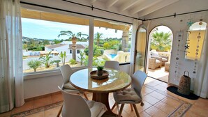 A dining area with a round table and chairs, large windows with curtains, and a view of a pool and greenery outside. - Golondrina - panoramic hillside holiday house in Moraira (Moraira)