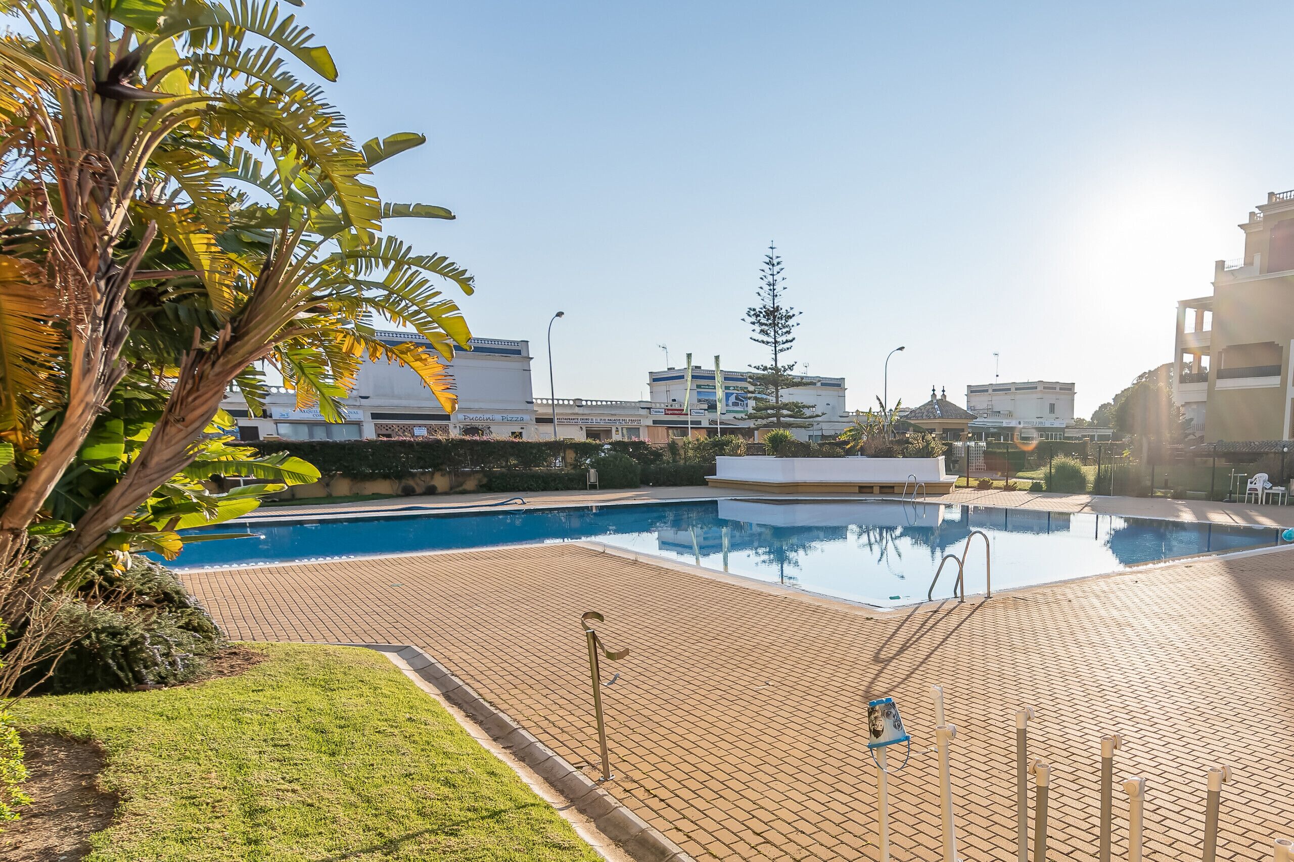 A swimming pool with a tiled deck, surrounded by greenery and buildings in the background.
