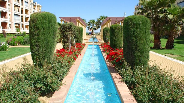 A pool with a water fountain, surrounded by neatly trimmed hedges and red flowers, with buildings in the background. - Canela Park Penthouse - Isla Canela Beach ICHP249P183 (Isla Canela)