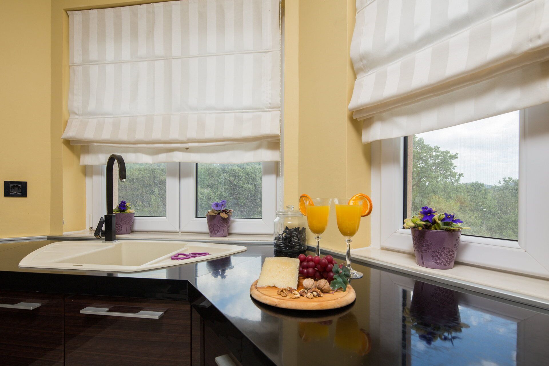A kitchen with a sink, two windows with white curtains, a countertop with a cheese platter, and two glasses of orange juice.