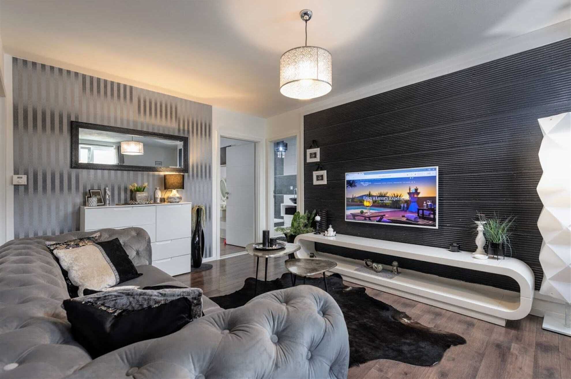Uma sala de estar moderna com uma grande TV de tela plana presa a uma parede preta, um sofá cinza, uma pequena mesa de centro redonda e um tapete preto.