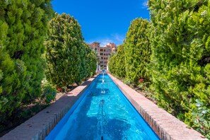 Ein langer, schmaler Swimmingpool, gesäumt von dichtem Grün, der zu einem mehrstöckigen Gebäude führt, über dem ein klarer, blauer Himmel erstrahlt.