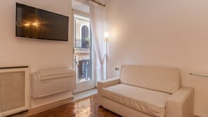 Una habitación con una televisión de pantalla plana montada en la pared, un sofá blanco, un radiador y una ventana con vistas a un balcón.