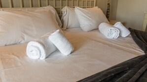 A neatly made bed with white linens and rolled towels, a wooden headboard, and a bedside table with a lamp.