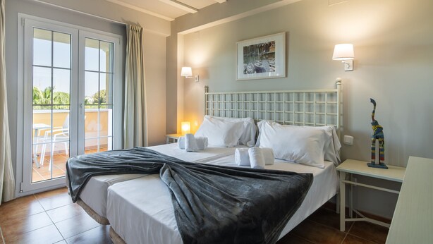 A hotel room with a bed, a nightstand, a lamp, a painting, and a view of the outdoors through a sliding door.
