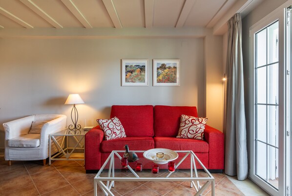 Una sala de estar con un sofá rojo, un sillón blanco, una mesa de centro de vidrio y dos cuadros enmarcados en la pared.
