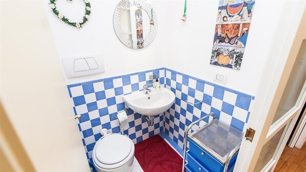 Een kleine badkamer met een witte wastafel, blauw-witte geruite tegels, een wit toilet, een rode mat, een spiegel en een decoratie aan de muur.