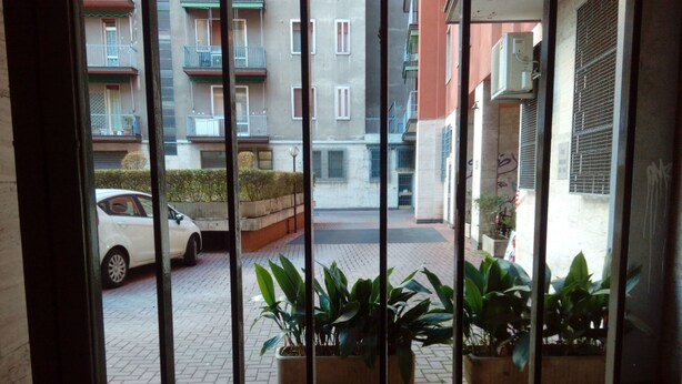 A view through a metal gate of a courtyard with potted plants, a white car, and a building with balconies and windows. - Casa Bella Fair (Bologna)