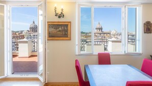 A room with a dining table and chairs, a painting on the wall, and large windows offering a view of a cityscape with domed buildings. - Argentina Penthouse in the heart of the historic center (Rome)