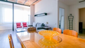 A spacious living area with a wooden dining table, red chairs, a grey sofa, and a red armchair. There is a bookshelf with books and a lamp on a side table.