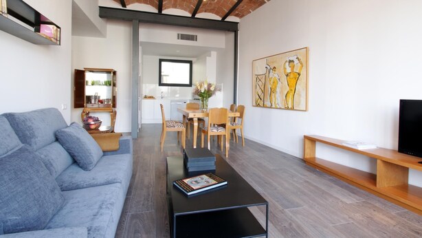 A modern living room with a grey sofa, a coffee table with a book, a dining area with wooden chairs, and a kitchen in the background.