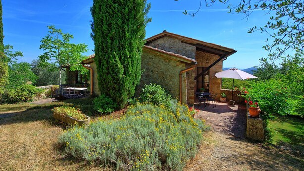 Una casa de piedra rústica con un porche cubierto, rodeada de vegetación exuberante y un cielo azul despejado.