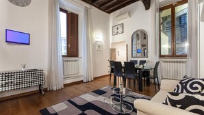 Een woonkamer met een bank, een salontafel, een televisie, een eethoek met stoelen en een tafel, en een raam met gordijnen.