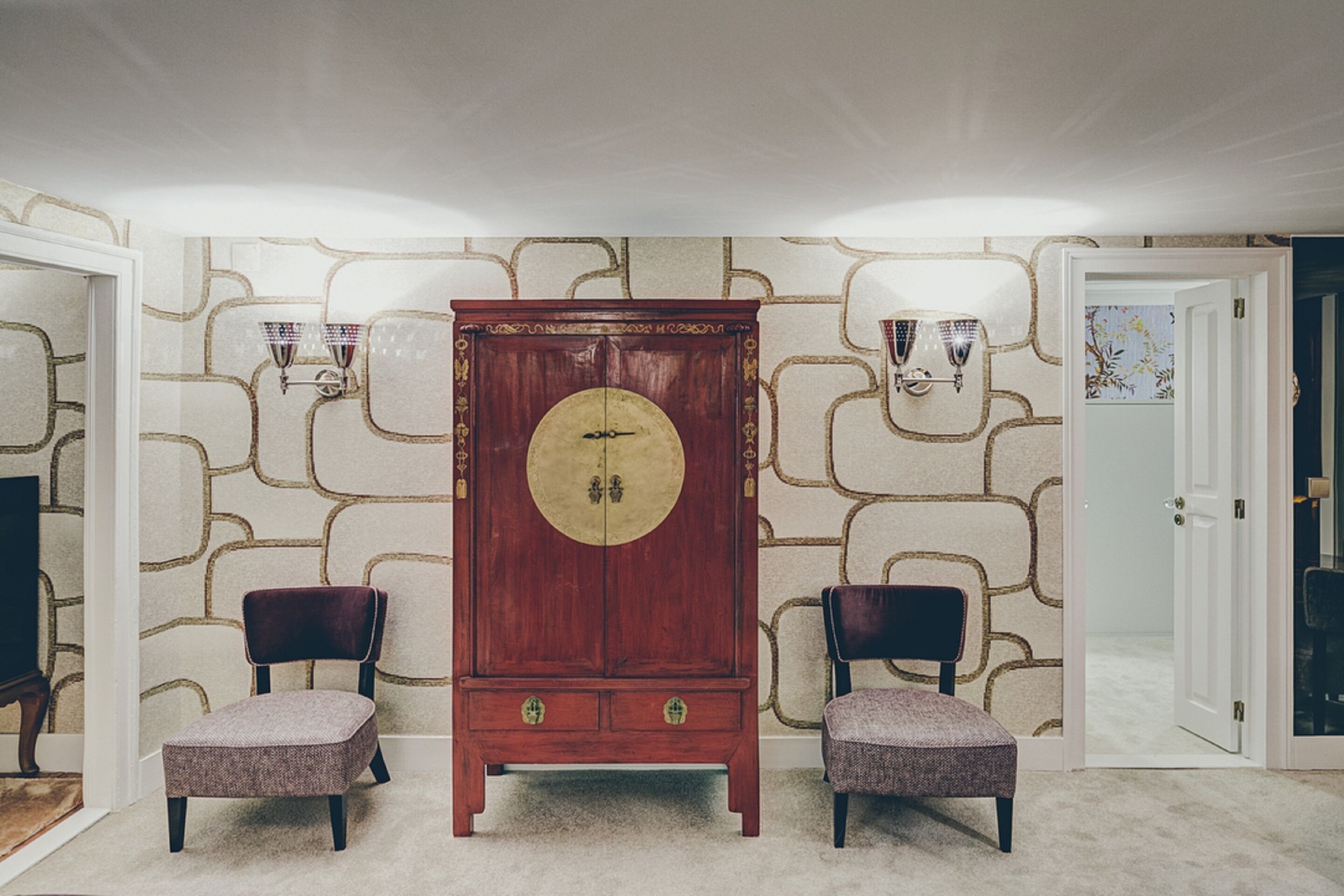 Ein Zimmer mit einem großen, hölzernen Schrank, zwei Polsterstühlen und Wandtapete mit Muster.