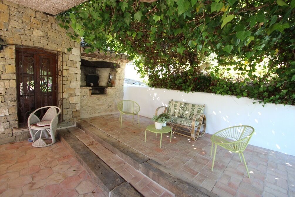 A stone building with a wooden door, a stone fireplace, and a white wall covered with ivy. There are three green chairs and a bench with cushions.