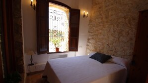 A bedroom with a stone wall, a bed with a white bedspread, a window with a metal gate, and a potted plant on the windowsill. - Rocasol - rustic finca for nature lovers in Benissa (Benissa)