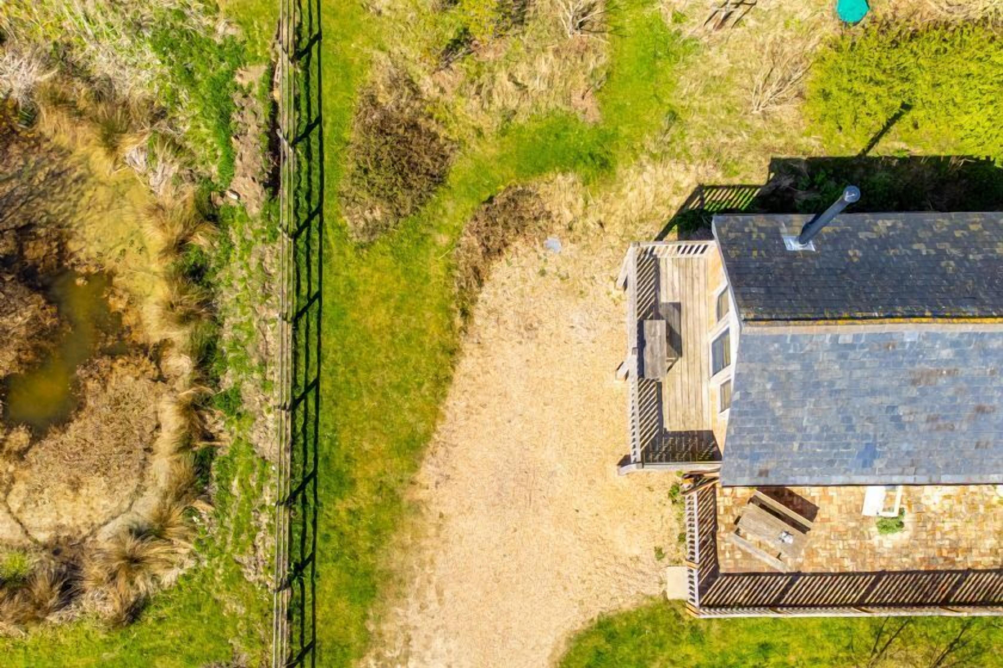 An aerial view of a house with a sloped roof, a fenced yard, and a small pond.