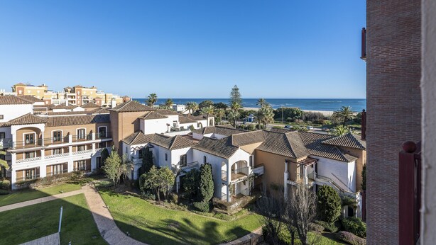 Uma área residencial costeira com edifícios que exibem telhados de terracota e varandas, um céu azul límpido e vista para o mar.