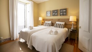 Una camera d'albergo con due letti, un'ampia finestra con tende, un comodino con una lampada e opere d'arte alle pareti.