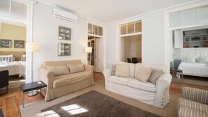 Un ampio soggiorno con due divani beige, un tavolino e una poltrona a righe. Alle pareti si notano quadri incorniciati e una grande finestra che lascia entrare luce naturale.