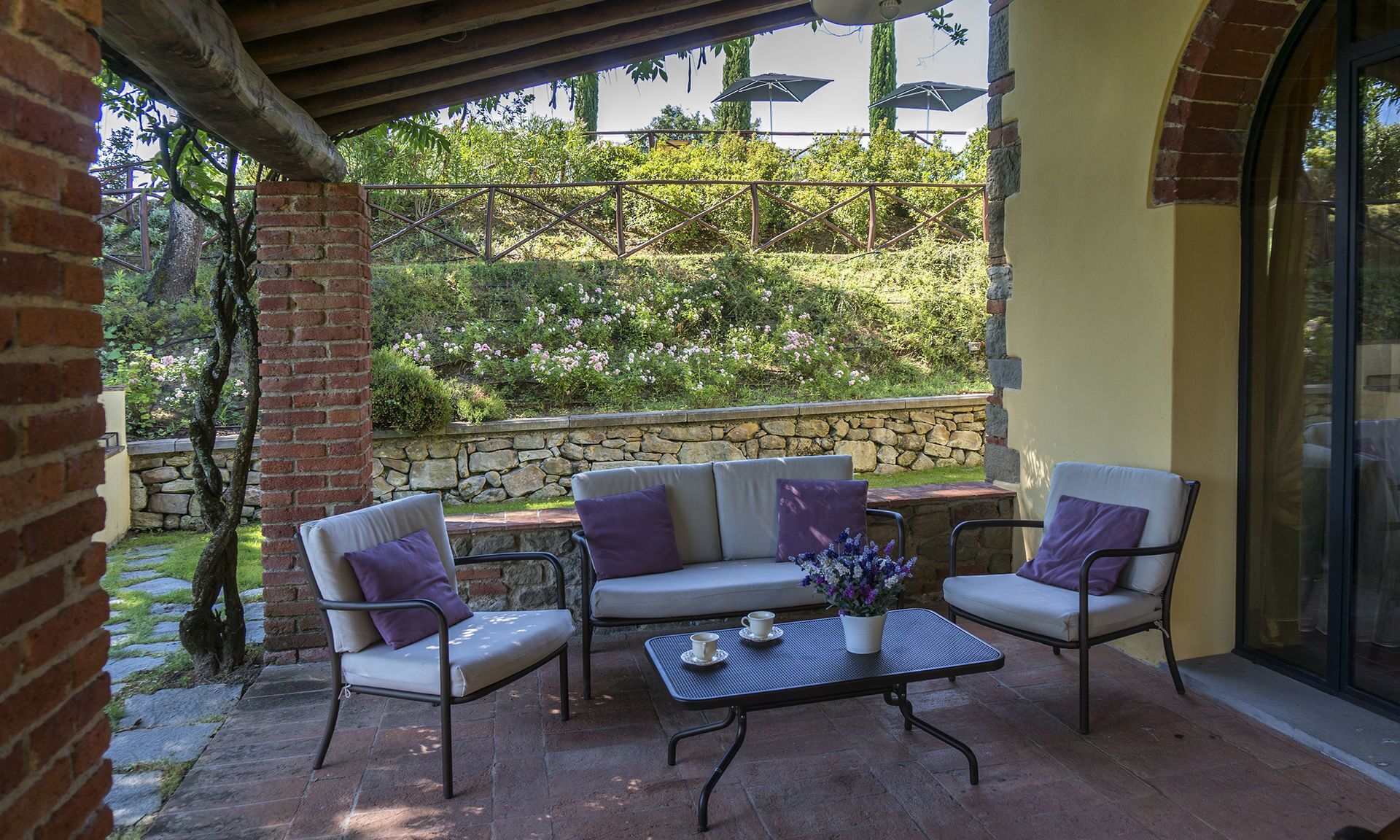A covered patio with a stone wall, a brick column, and a view of greenery.
