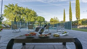 Zona d'esmorzar a l'aire lliure amb una taula preparada per a menjar, rodejada de cadires, i vistes a un jardí amb arbres alts.