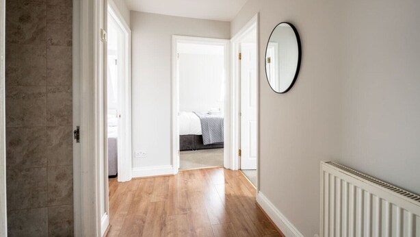 A hallway with a wooden floor, white walls, and a round mirror on the right. - Comfy 2 Bed Flat (QCE) (Greater London)