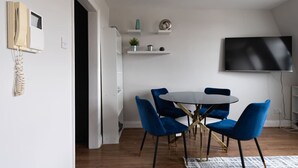 A modern dining area with a round table and blue chairs, a flat-screen TV mounted on the wall, and a small shelf with decorative items. - Comfy 2 Bed Flat (QCE) (Greater London)