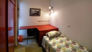 Une petite pièce comprenant un lit, un bureau, une chaise, une lampe fixée au mur et un tableau accroché au mur.