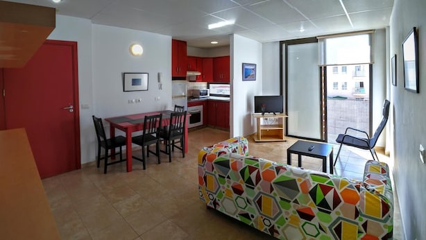 Un salon avec un canapé aux couleurs vives, un coin repas avec une table et des chaises rouges, une cuisine avec des placards rouges et un balcon offrant une vue dégagée.