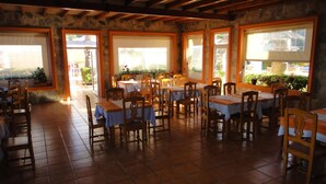 Un comedor interior con varias mesas y sillas, paredes de piedra y ventanas amplias que permiten la entrada de luz natural.