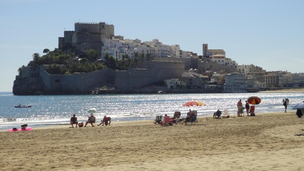 Una playa con gente relajándose bajo sombrillas, un complejo de edificios históricos al fondo y un cielo azul despejado.