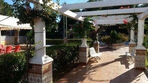 Un área de patio techada con piso de baldosas, columnas blancas con bases decoradas y sillas de plástico rojas.