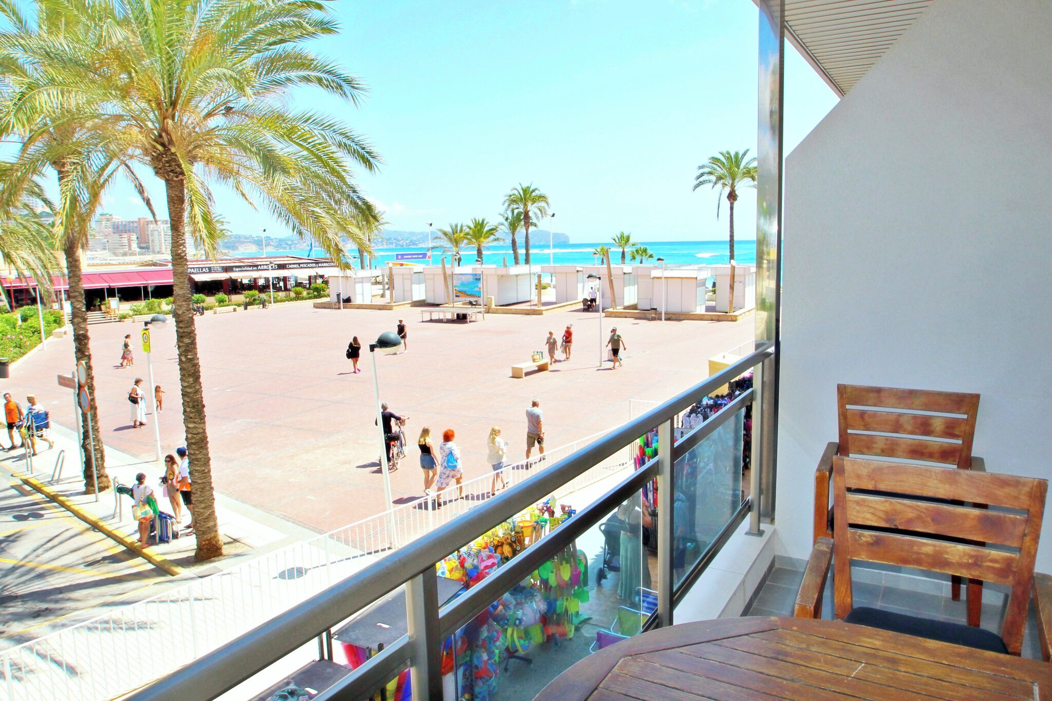 A balcony with wooden chairs and a view of a beach with palm trees and people.