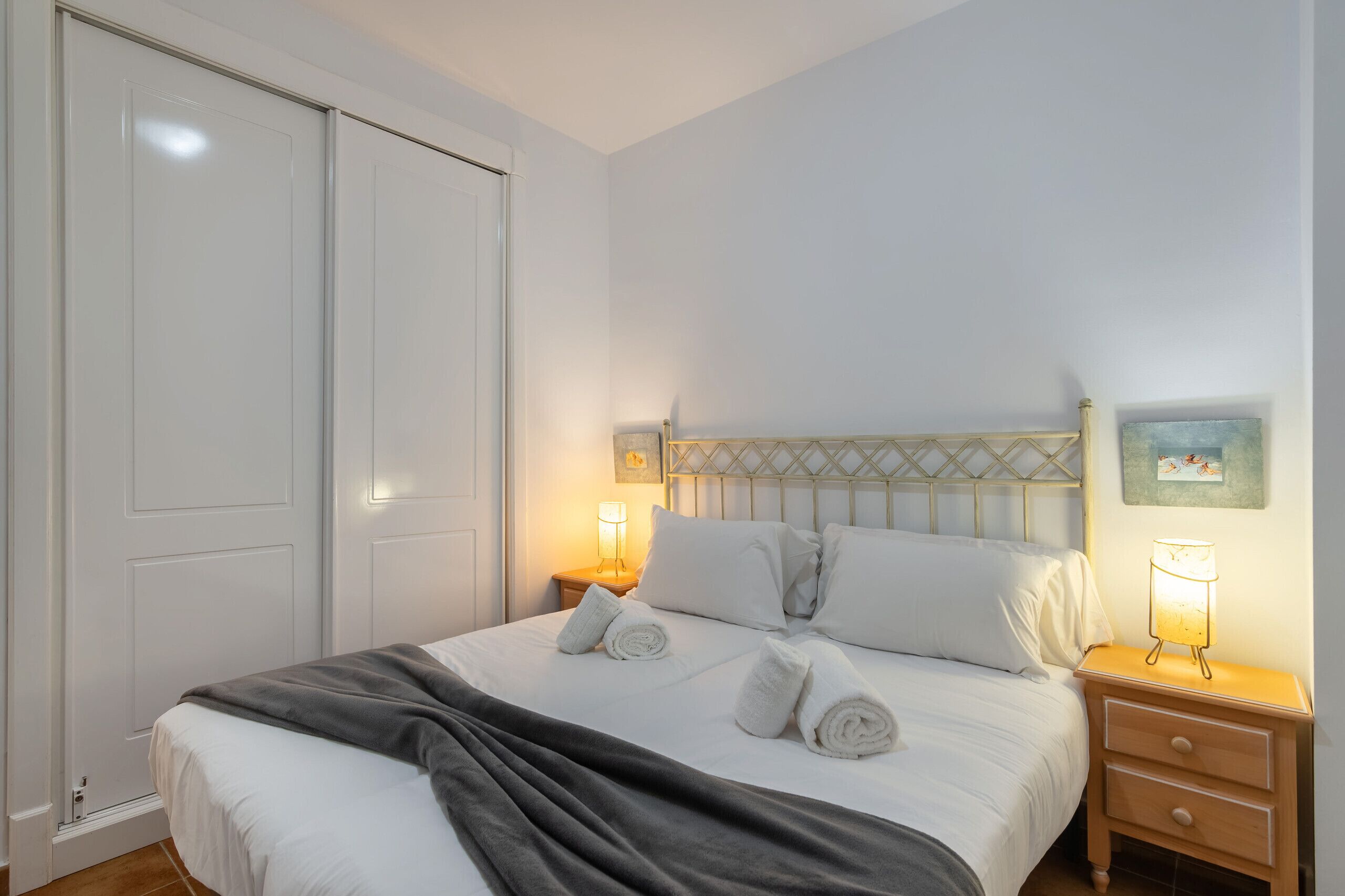 Una cama bien hecha con sábanas blancas, un plaid gris y dos toallas enrolladas. Hay dos lámparas de noche sobre mesitas de madera.