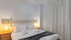 Een net opgemaakt bed met witte lakens en handdoeken, een houten hoofdbord, bedlampjes en een gordijn.
