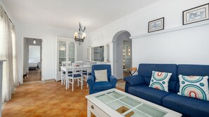 Un salon avec un canapé bleu, une table basse en verre et un coin repas avec des chaises blanches.