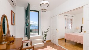 A room with a wooden desk, a chair, a mirror, a bed with a pink bedspread, and a view of the sea through an open door. - Villa Brač Stone with Swimming Pool (Bol)