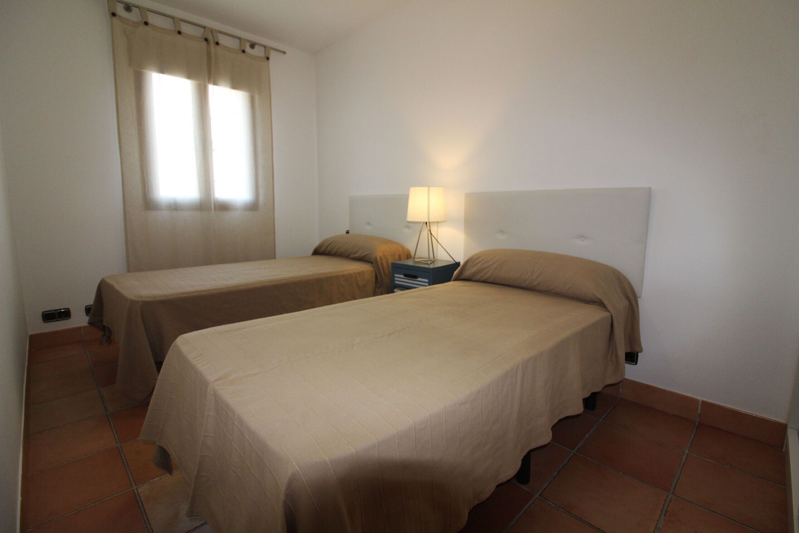 A room with two single beds, a nightstand with a lamp, and a window with curtains.
