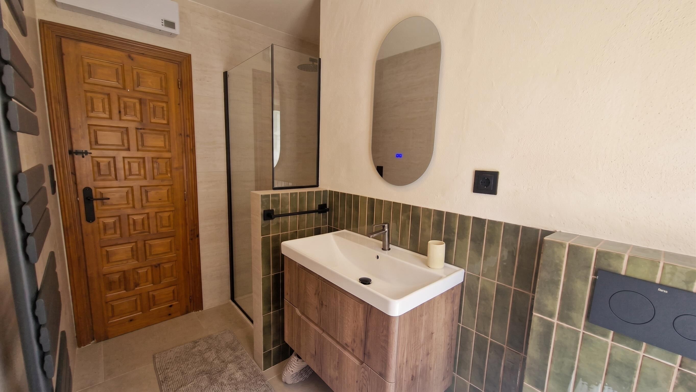 A bathroom with a wooden door, a shower, a sink with a mirror, and a tiled wall.