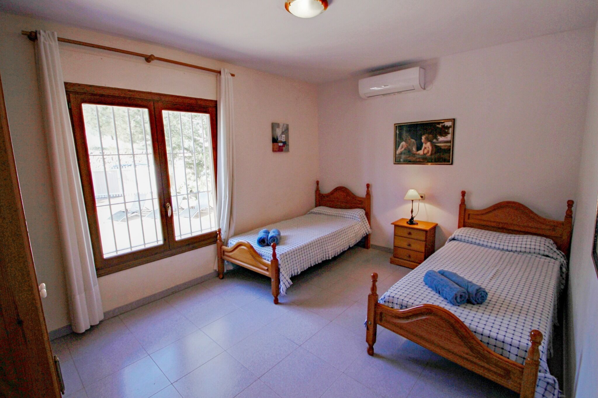 Un dormitorio con dos camas individuales, una mesita de noche con lámpara, una ventana con cortinas y un aire acondicionado instalado en la pared.