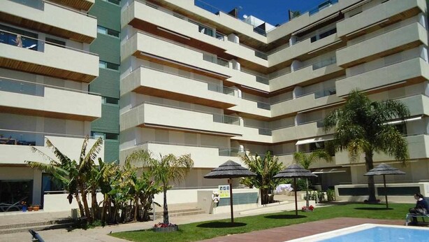 A multi-story hotel with a swimming pool, outdoor seating, and palm trees. - Luxury apartment in private condominium, with pool (Quarteira)